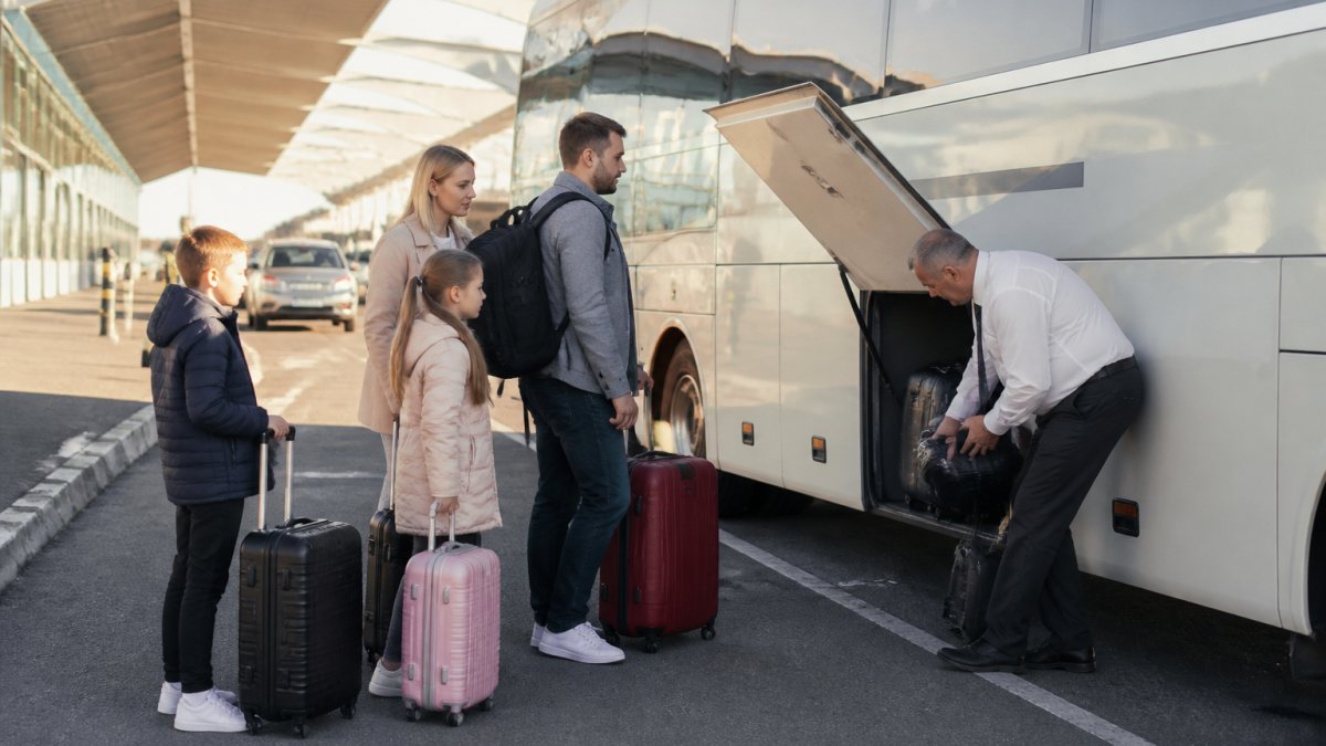 A family with suitcases boarding a shared coach outside an airport terminal, driver helping load luggage into the hold, morning light